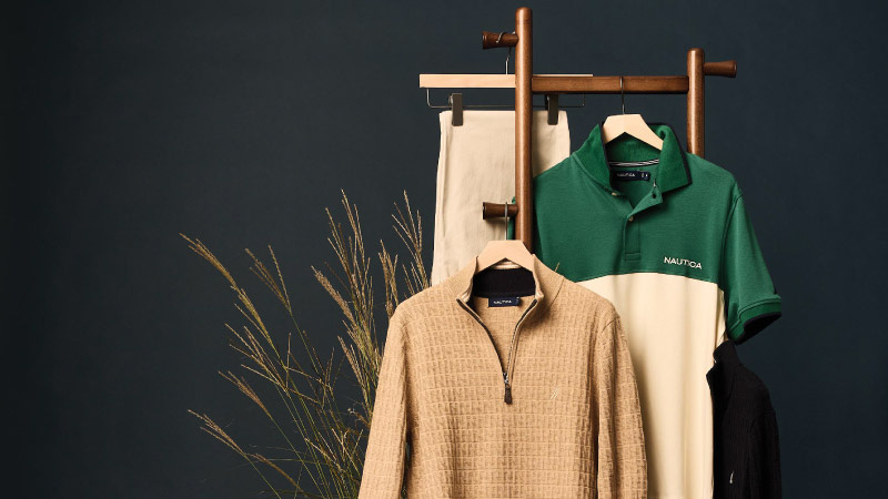 Clothing display against a dark backdrop shows beige pants, a beige textured zipper sweater, and a green and cream polo. Decorative plants add a natural touch.