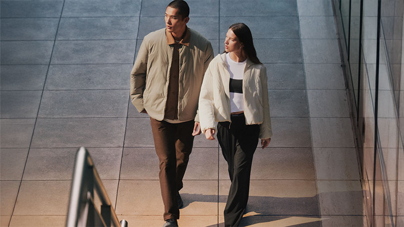 A man and woman walk confidently on a sunlit urban walkway. Both wear light jackets with relaxed expressions, exuding a calm, stylish vibe.