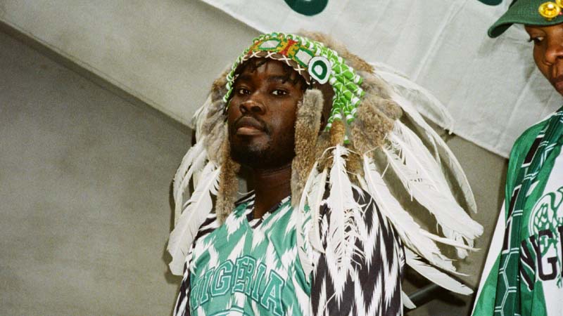 A person wears a Nigeria jersey and a feathered headdress with green decorations, appearing proud and celebratory. 