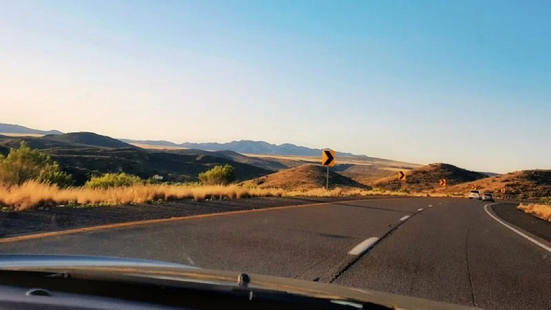 A view from a car driving on a curved highway through dry, rolling hills under a clear sky, with road signs visible ahead.